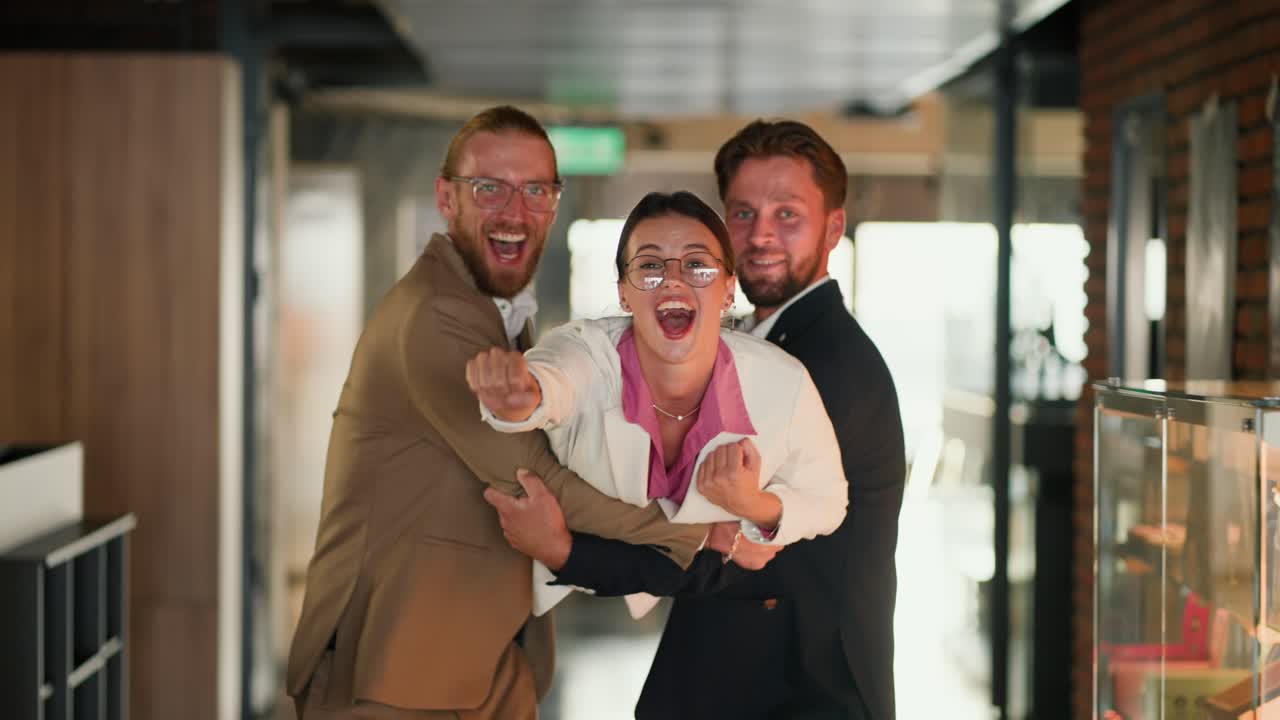A blond man in a brown suit and a brunette man in a blue jacket carry in their arms a brunette girl in a pink shirt and white jacket. Playing Superman in the office. The girl extended her hand forward and was carried by two guys