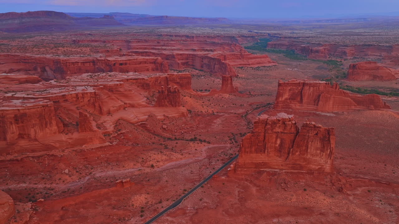 Fantastiske røde steiner med bratte skråninger og flate topper. En motorvei krysser det øde landskapet i Arches nasjonalpark i Utah, USA ved solnedgang. Luftperspektiv
