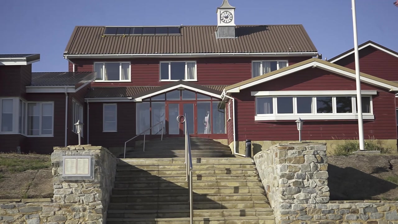 Sollys belyser en rød bygning med et fremtredende ur og steintrapper, og skaper en slående arkitektonisk scene. Strukturen står mot en klar blå himmel.