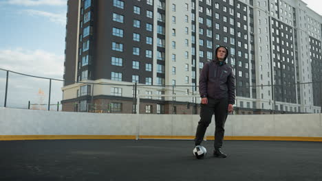 deportista retorciendo hábilmente los tobillos de la pierna derecha y izquierda con una pelota de fútbol delante en una moderna arena deportiva urbana, con altos edificios de oficinas y postes de portería visibles en el fondo