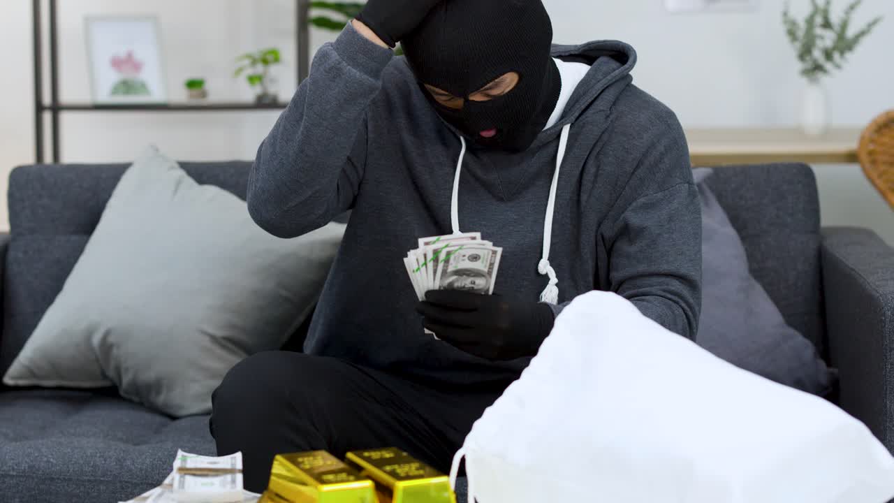 A man in a balaclava counts money and gold bars before revealing his face