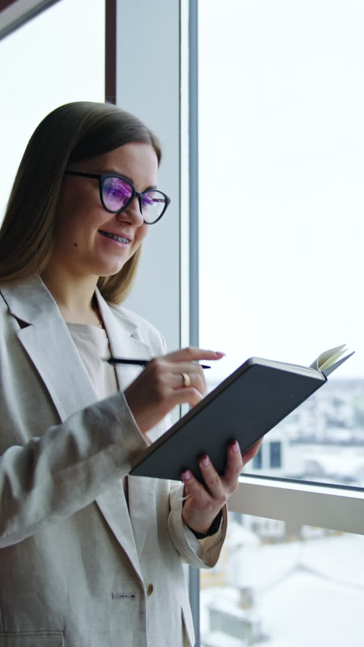 Smiling young woman writing something down in her book. Taking notes while standing a t the window. Cityscape in blur at the backdrop. Vertical video