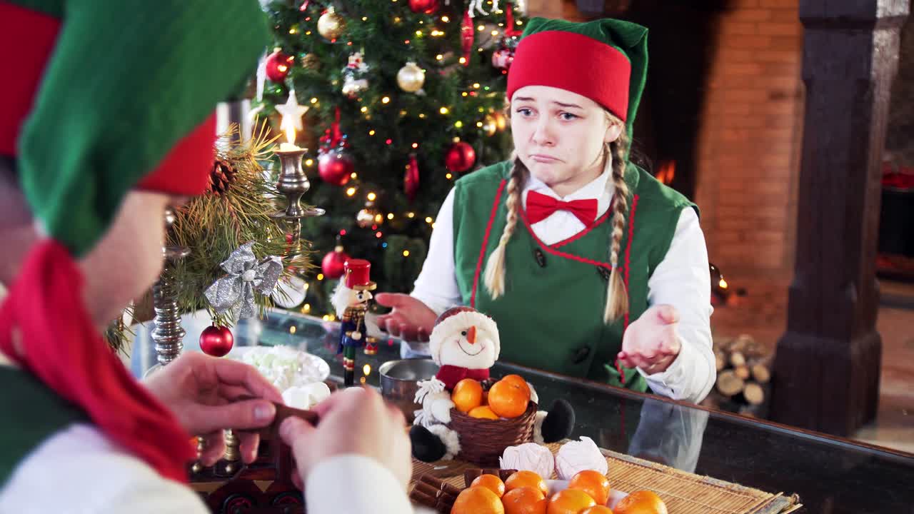Two funny elves sitting at table on Christmas background. Girl elf tries to take sweets and another elf interferes her