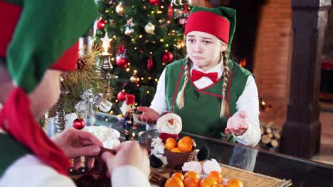 two funny elves sitting at table on christmas background. girl elf tries to take sweets and another elf interferes her