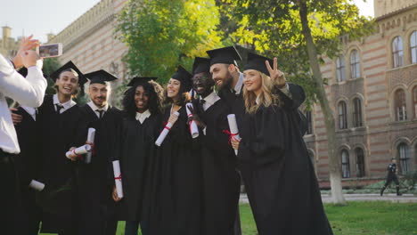 Vista-Trasera-Del-Hombre-De-Cabello-Gris-Tomando-Una-Foto-En-El-Teléfono-Inteligente-Del-Grupo-De-Graduados-Multiétnicos-En-Vestidos-Negros-Tradicionales-Y-Gorras-Con-Diplomas-En-Las-Manos