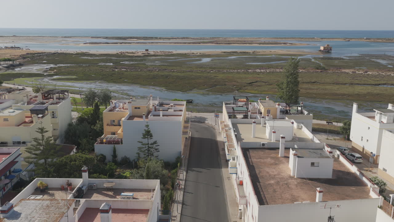 Drone moving forward above a quiet street in Fuzeta, showing rooftops, trees, cars, tidal flats, and the distant rescue station