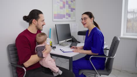 una confiada chica morena con gafas y un uniforme azul el médico da consejos sobre el desarrollo adecuado del niño en una recepción con un joven hombre moreno padre junto con su pequeña hija bebé en una clínica moderna