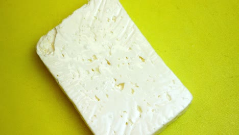 a hand places down a block of feta or greek style salad cheese onto a yellow counter or chopping board