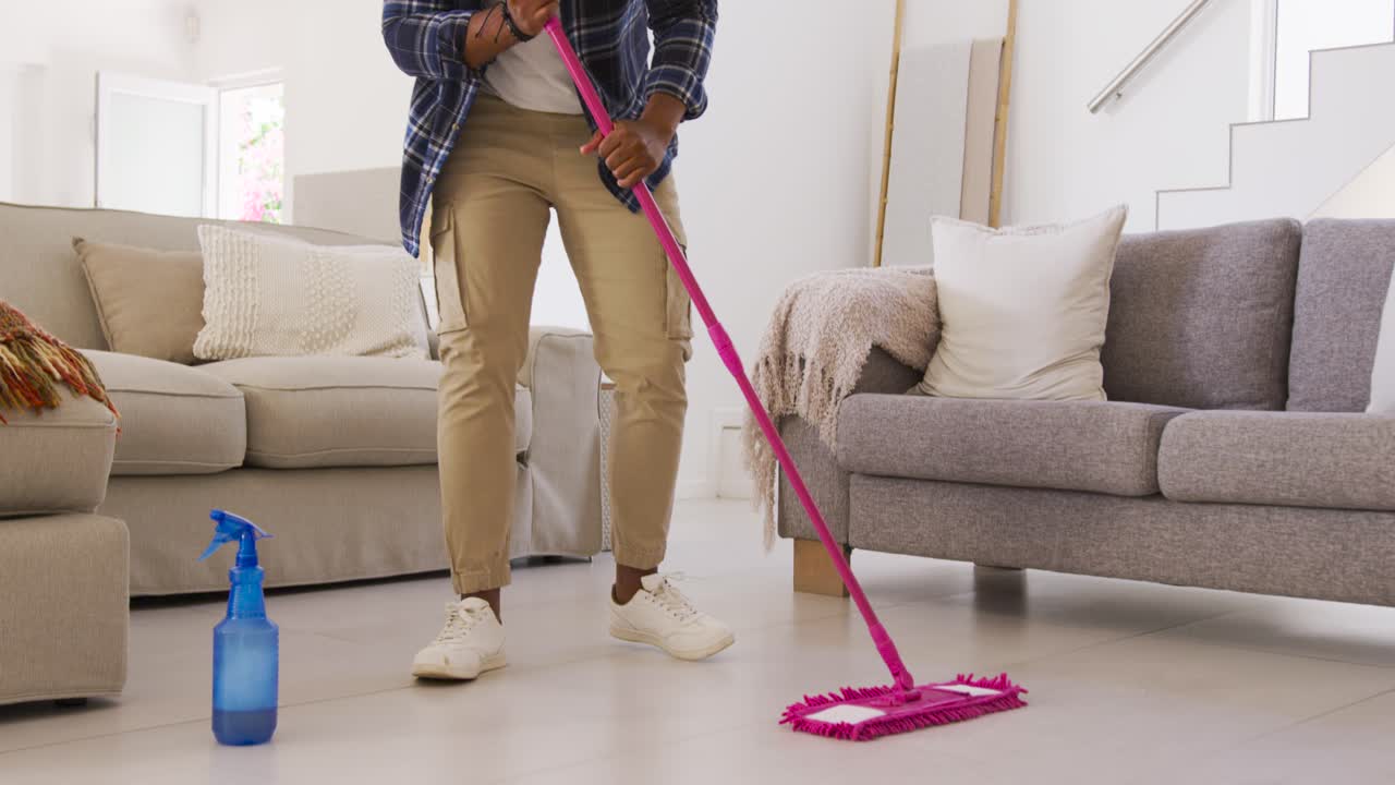 Afroamerikansk mann forbereder blå sprayflaske, mopper flisgulv med rosa mopp for rengjøring.
