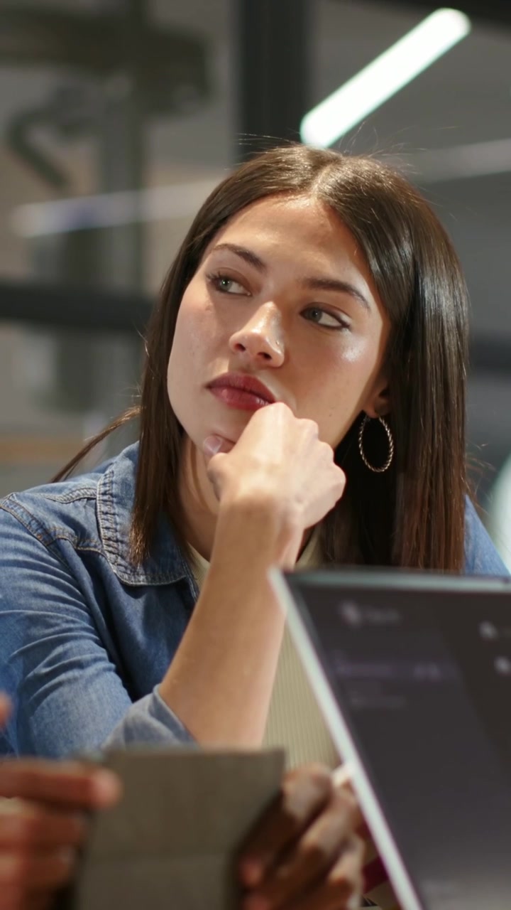 Video verticale: Donna in denim segue un collega che mostra un tablet in ufficio, controllando documenti sul laptop.