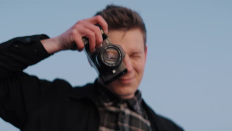 joven fotógrafo caucásico tomando fotos con una cámara fotográfica analógica durante la puesta de sol contra el cielo azul