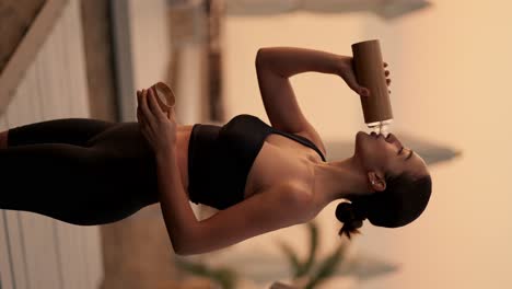 vertical video: a brunette girl with tied hair in a black sports uniform drinks water from a sports bottle on a sunny beach with palm trees