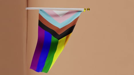 progress pride flag waving on pole against solid yellow background. fabric texture with rainbow colors and chevron stripes in blue, pink, white, brown, and black. lgbtq+ identity and equality vertical