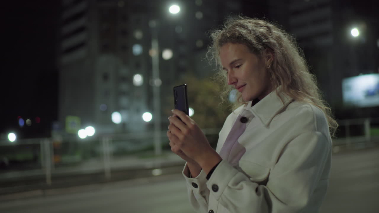 Young woman in white coat focused on smartphone while standing near quiet road at night, with passing car and glowing street lights illuminating urban backdrop in soft ambient bokeh lighting