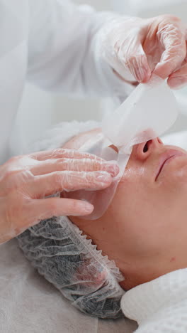 cosmetologist applying regeneration moisturizing sheet mask on young adult woman face skin and eyes