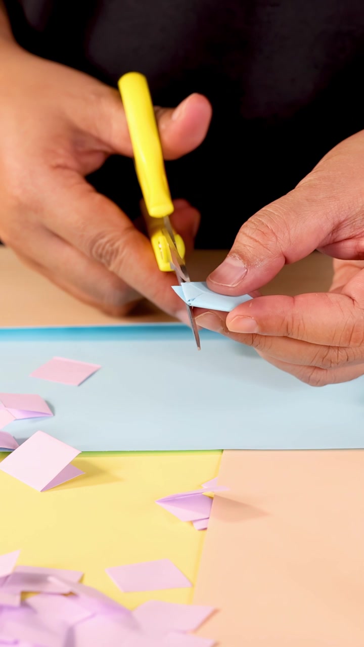 Person uses scissors to cut and fold pastel paper squares on a table, close-up view