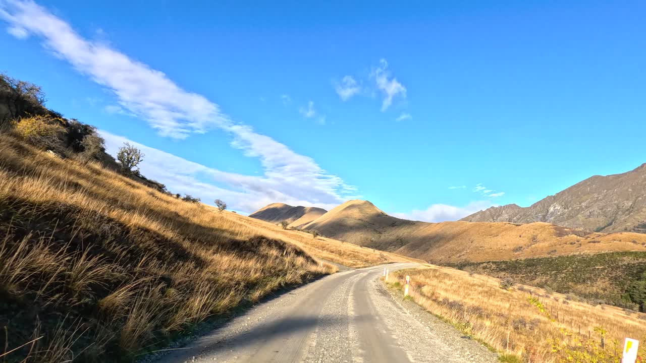 Vehicle drives along gravel mountain road, golden hills, clear sky, bright daylight, smooth timelapse motion