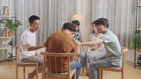 asian teen group sitting in chairs forming a circle discussing at home. teen students support a boy in brown t-shirt not to crying