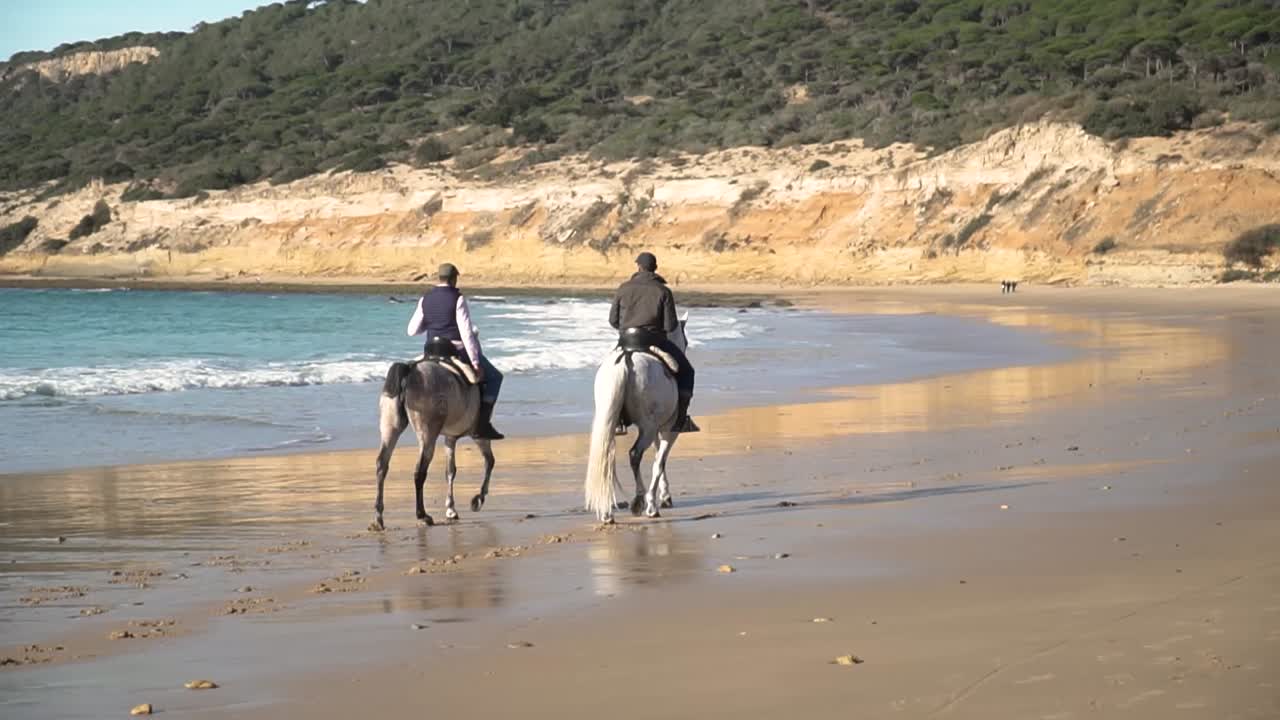 amplia toma manual de dos jinetes con sus caballos galopando a lo largo de la playa con olas tranquilas que dominan el paisaje
