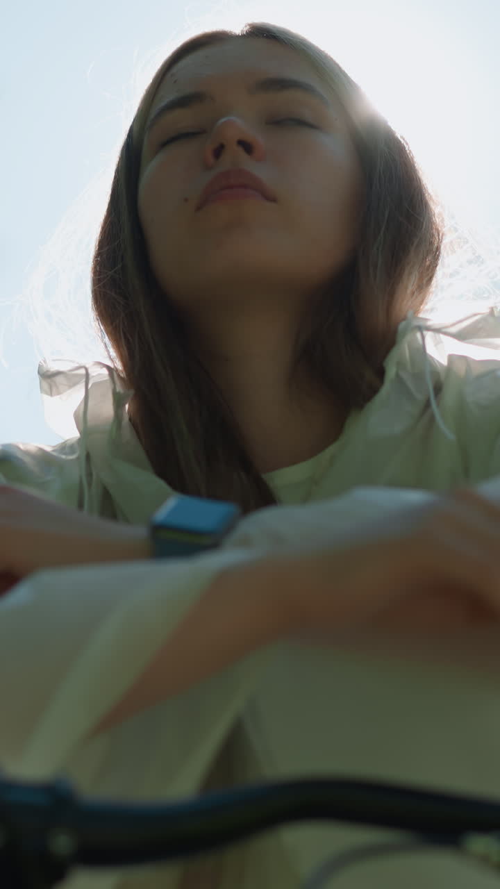 반투명한 레인코트를 입은 여성이 밝은 빛에서 후드를 내려 놓고, 그녀의 상하는 얼굴 주변에 부드럽고 에테리얼한 빛을 만들어, 배경에는 흐릿한 초록색이 있습니다.