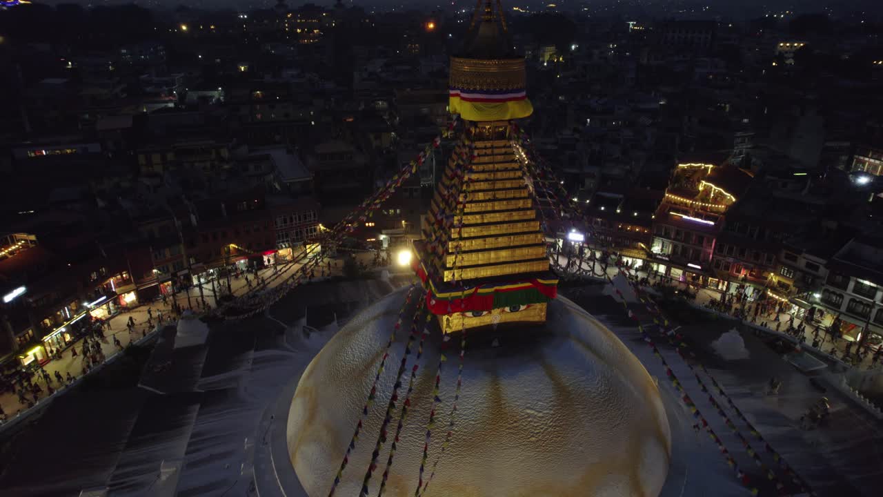 A calm night drone view of Boudhanath Stupa, with boudha and nath noted separately, glowing above Kathmandu. The sacred Buddhist landmark stands radiant in the quiet evening cityscape