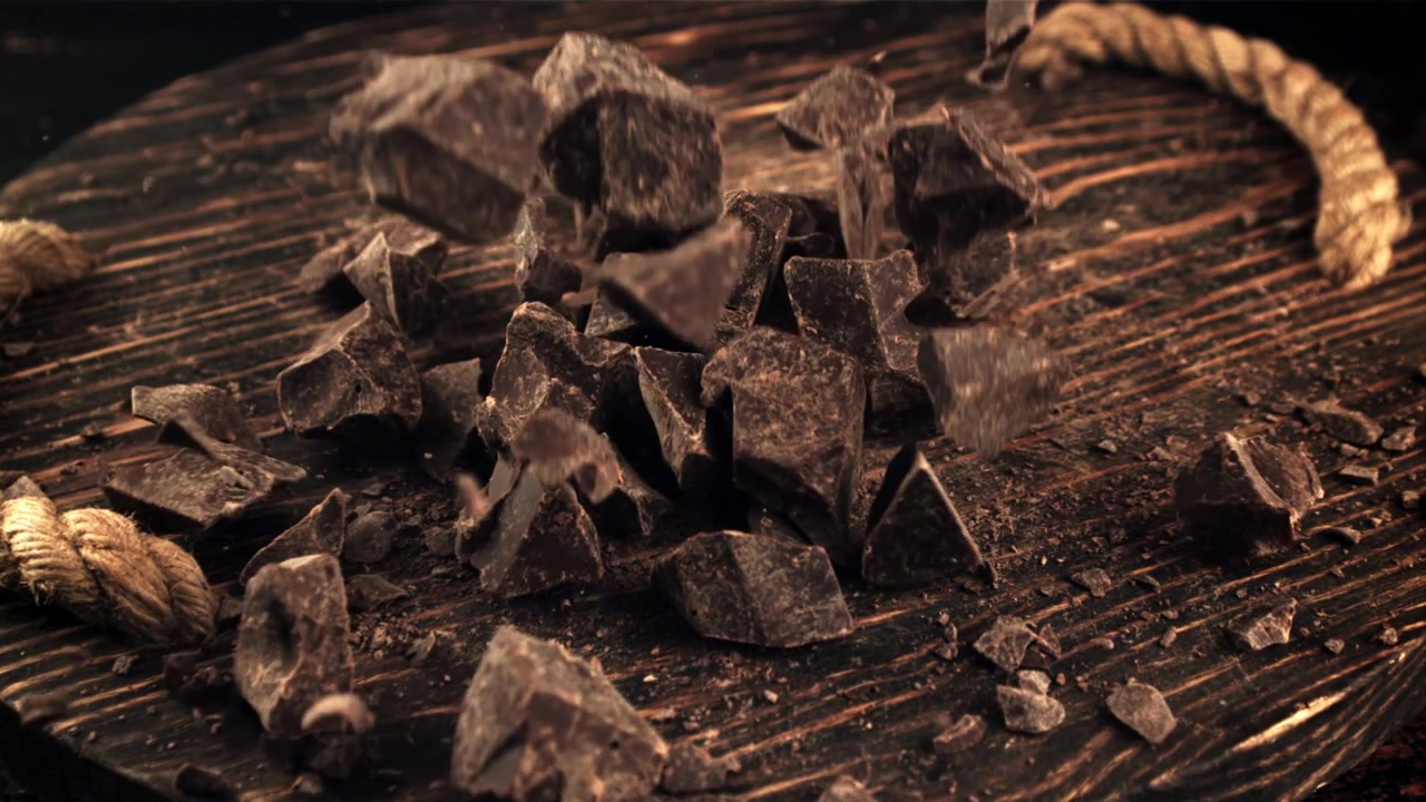 Pezzi di cioccolato fondente che cadono al rallentatore su un tagliere di legno rustico.