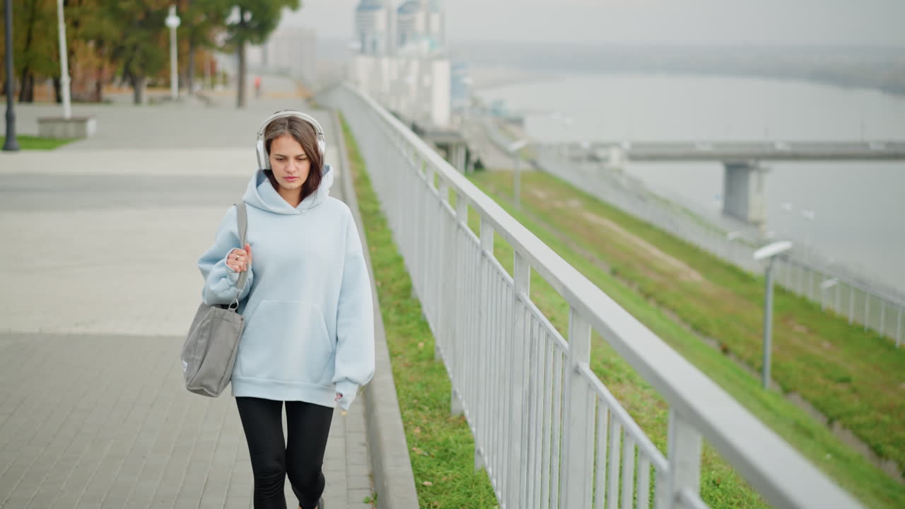 Pretty woman holding ash bag walking beside iron rail, with blurred view of overhead bridge and road in background, peaceful urban outdoor setting, casual fashion, strolling with confidence