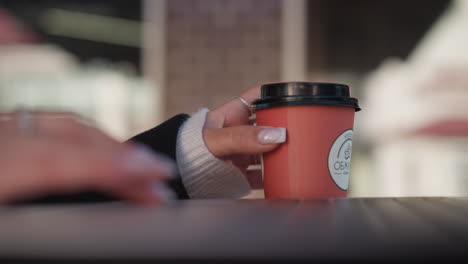 close-up of lady's hand gently turning coffee cup near laptop with focus on manicured fingers, blurred typing hand, and chair in background