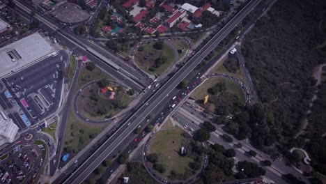 Cloverleaf-intersection-in-the-south-of-Mexico-City,-where-several-cars-are-circulating,-a-parking-lot,-soccer-fields-on-the-lower-right-corner,-trees-and-some-red-roofs