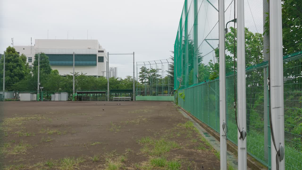 An empty, wide shot of an urban baseball field from the side, showing the fence and dugout area