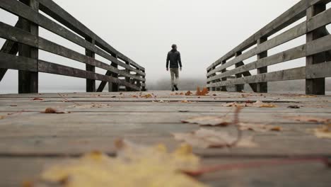 Hombre-Caminando-En-Un-Muelle-De-Madera-Con-Hojas-De-Color-De-Otoño-En-El-Suelo