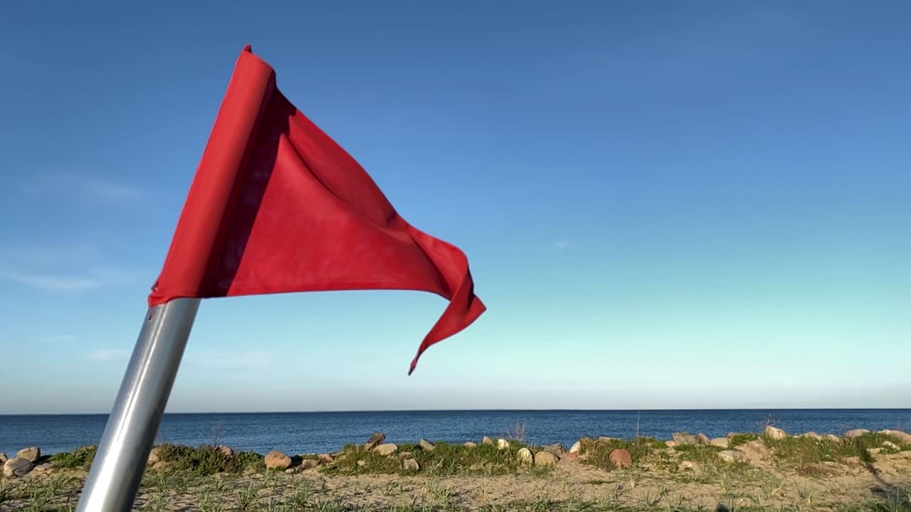 Red Warning Flag On Pole Near Coastline Waving In Wind Against Sea And Blue Skies. Slow Motion