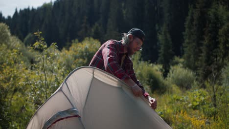 A-blond-man-in-a-red-plaid-shirt-set-up-a-tent-in-a-clearing-near-the-mountains