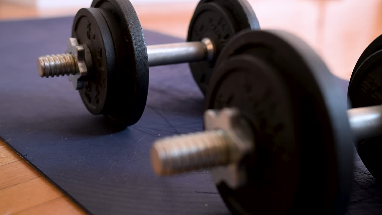 A rack focus shot shifts from the front dumbbell to the back one. A cinematic close-up of free weights on a yoga mat, ready for a home workout or strength training