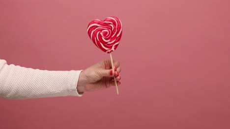 profile side view closeup of woman hand holding, showing red lollipop in heart shape turning candy.