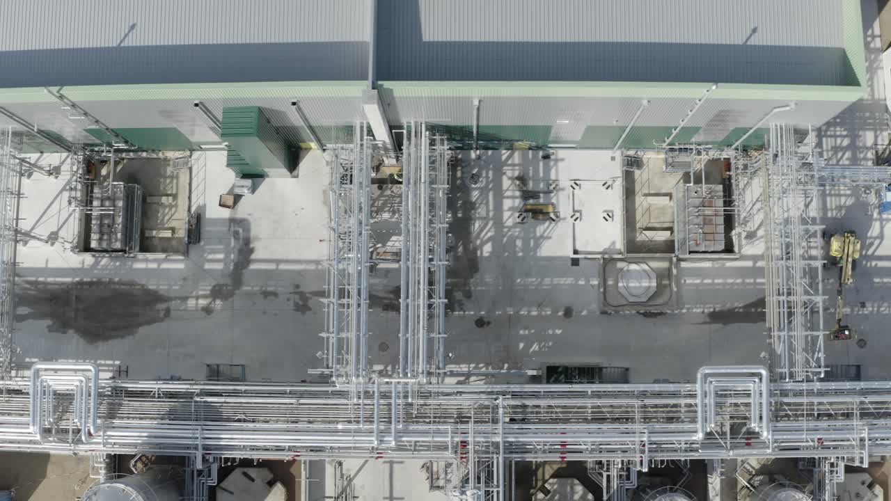 Top-down aerial tracking over complex network of pipes and industrial equipment on factory rooftop, workers visible, Castets, France