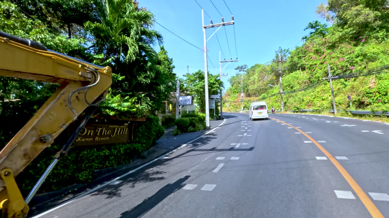 Excavator and cars drive uphill on a tropical road with lush greenery in bright daylight