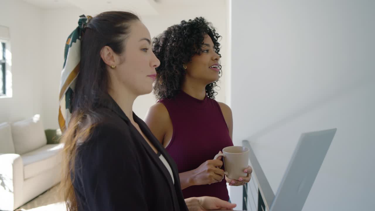 Diverse female agent and client opening laptop, holding mug while viewing home on landing
