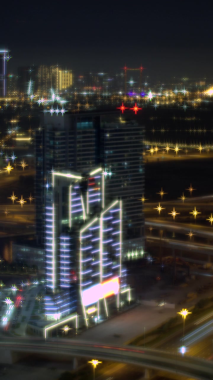 Timelapse vertical de um arranha-céu moderno em Dubai iluminado por luzes neon brilhantes à noite, com rastros de luz do tráfego da rodovia abaixo.