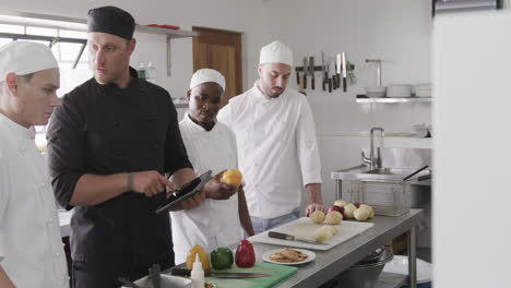 diverse male chef instructing group of trainee male chefs using tablet in kitchen, slow motion