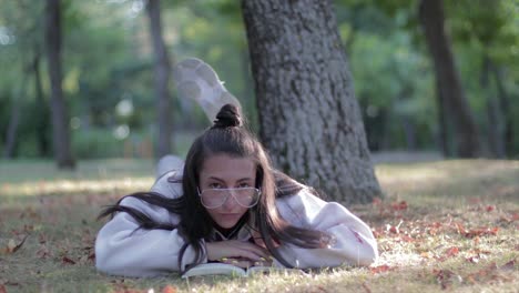 Chica-Con-Grandes-Anteojos-Tumbada-En-El-Césped-Leyendo-Un-Libro-En-Un-Parque