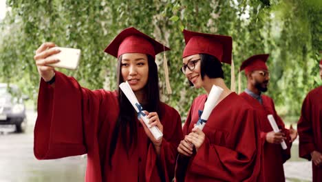 pretty girls graduating students are taking selfie with diploma scrolls using smartphone, women are posing and smiling, their fellow students are celebrating in background.