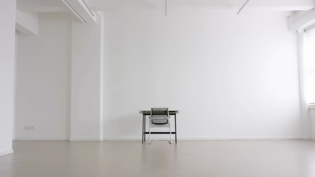 Spacious modern studio interior with centered desk and chair beside large white wall, exposed ceiling beams, soft natural daylight, clean neutral surfaces, and calm contemporary workspace mood