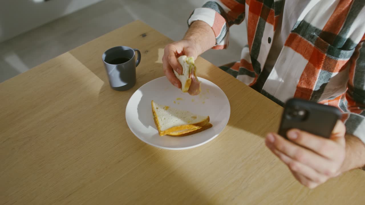 hombre comiendo sándwich y bebiendo café mientras usa un teléfono inteligente