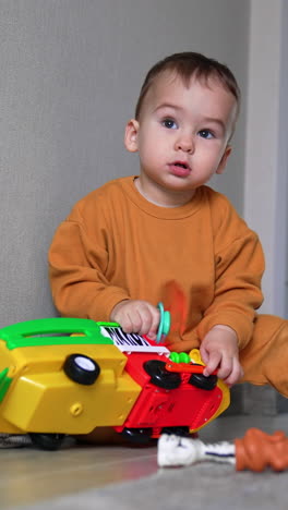 lovely calm baby boy playing with a bright lorry. child sits on the floor at the wall pressing buttons on a toy. black cat appears behind. vertical video
