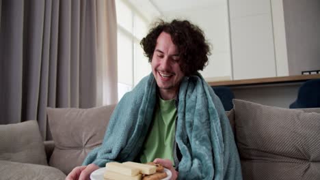 happy cheerful brunette guy with curly hair in a blue plaid actively laughs and eats sweet snacks while watching a funny movie and comedy at home in a modern apartment