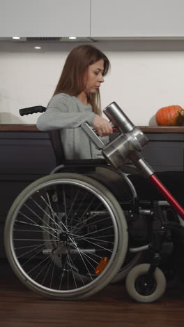 young woman in wheelchair takes care of house keeping clean. calm long-haired female with injury vacuums in kitchen using modern vacuum cleaner side view