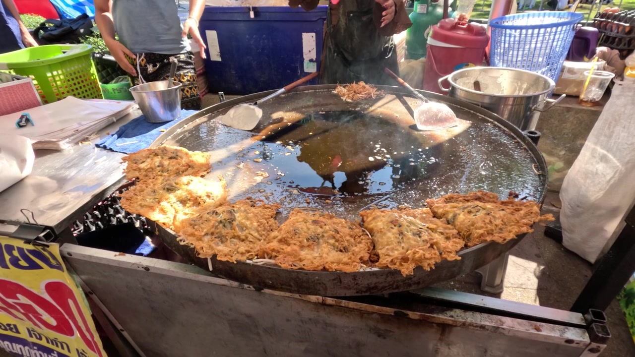 Potret jarak dekat seorang penjual kaki lima yang menggoreng omelet tiram renyah dalam minyak panas di luar ruangan.