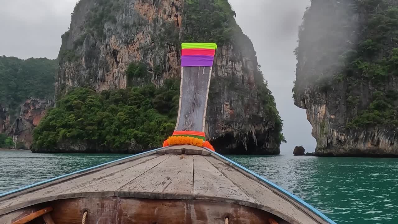 A longtail boat glides through emerald waters between towering limestone cliffs under an overcast sky.