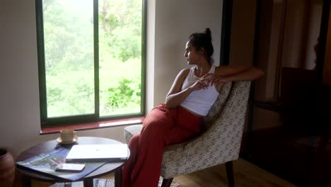 A-woman-in-a-white-tank-top-and-red-pants-sits-in-a-chair,-gazing-out-a-large-window